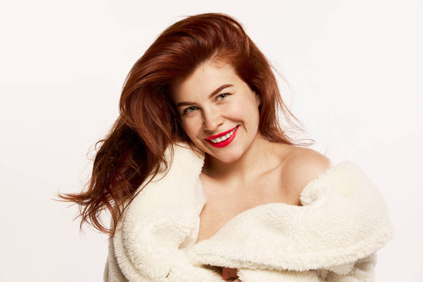 Portrait of young beautiful red-haired woman posing in fur coat, smiling isolated over white background. Teeth whitening, dental care. Concept of hight fashion, style, make-up, beauty, magazine style - Fotografie, Obrázek