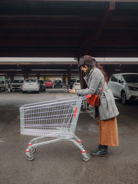 mujer adulta joven hispana empujando carrito leyendo la lista de compras de teléfonos inteligentes en el estacionamiento en la gran tienda de supermercados - Foto, imagen