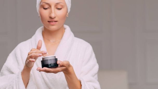 Young attractive woman in a bathrobe applies cream on her face in the bathroom. - Φωτογραφία, εικόνα
