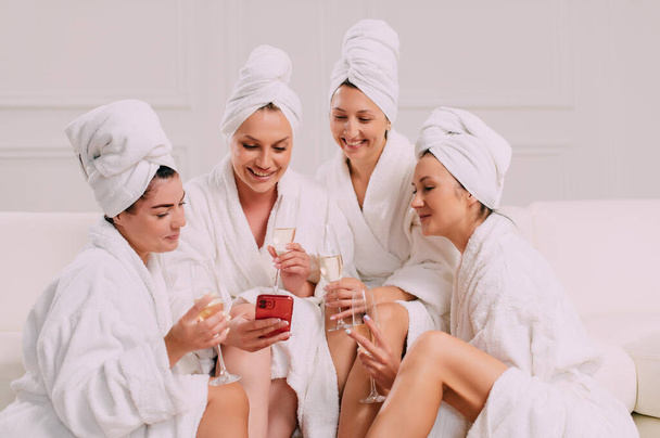 A group of women spend leisure time. Four Young attractive women in bathrobes look at the smartphone screen and smile. - Photo, image