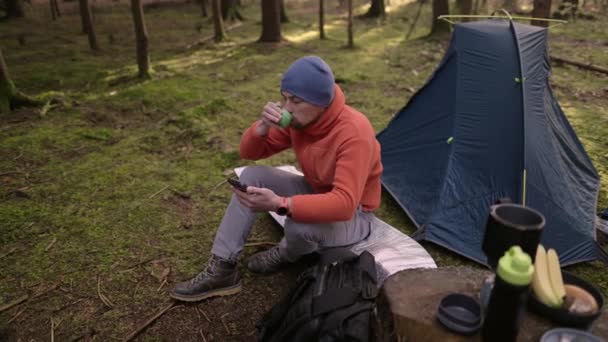 Morning at campground in fall forest. Tursit is browsing Internet on his smartphone while sitting on hiking mat near tent and drinking a beverage from a cup. Travel, people and healthy lifestyle.  - Footage, Video
