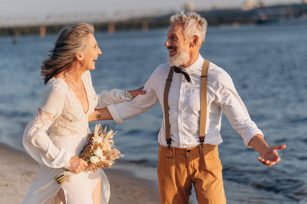 Cheerful elderly couple walkiing along beach holding hands. - Photo, image