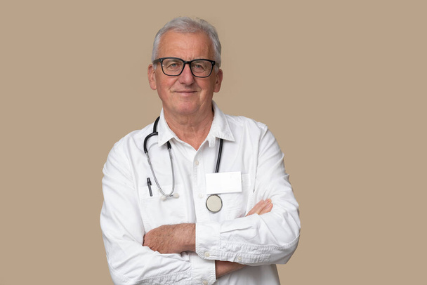 Starší lékař v brýlích se stetoskopem a bílou uniformou na světle hnědém pozadí. - Fotografie, Obrázek