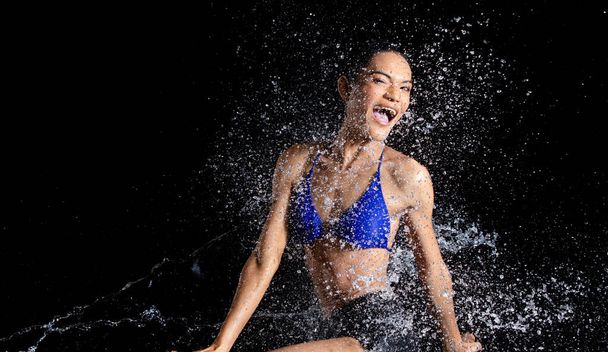 Gebräunte Haut Asiatin im Bikini posiert im Aqua-Studio. Spritzwassertropfen breiten sich auf den Körper aus. Spaß Emotionen weibliche Mädchen auf Wasser Angriff Flattern und Stop-Motion-Freeze-Schuss, schwarzer Hintergrund isoliert - Foto, Bild