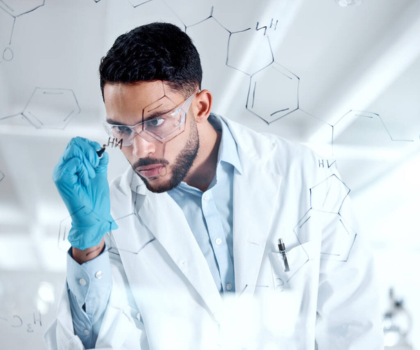 Young focused hispanic chemist drawing a dna structure on a board while brainstorming. Serious scientist planning his research experiment in the lab, wearing safety goggles. Young man in a lab. - Photo, Image