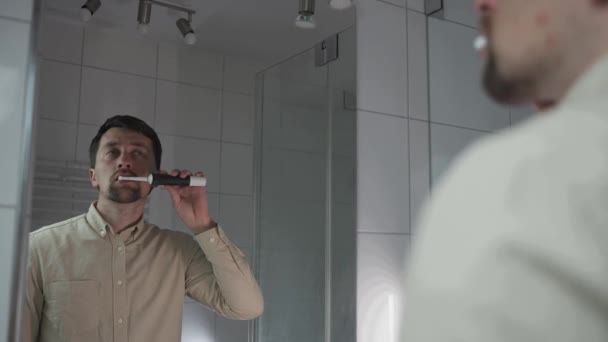 Homem caucasiano escova os dentes com uma escova de dentes elétrica no banheiro, vista do reflexo através do espelho. Procedimentos de higiene matinal. Irrigador oral. Rotina matinal de belo sorriso - Filmagem, Vídeo