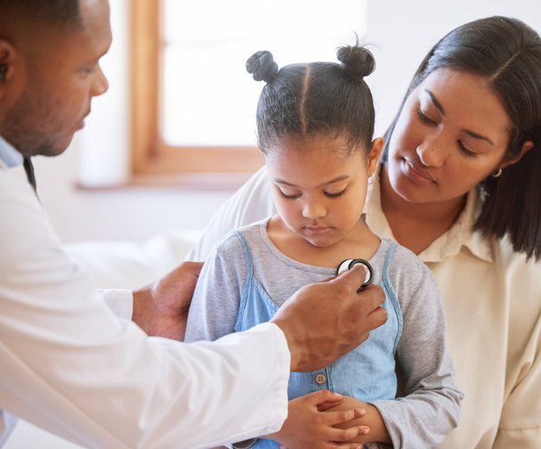 Sad little girl at doctors office. Sick girl sitting with mother while male paediatrician listen to chest heartbeat. Male doctor examining child with stethoscope. Mom holding kid during doctor visit. - Photo, Image
