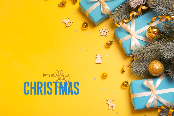 Merry Christmas text on yellow background with blue gifts, pine tree and festive decorations top view. Xmas greeting card. - Photo, Image