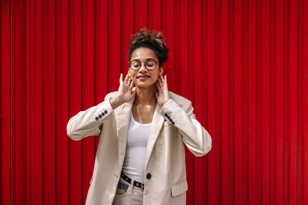 Позитивна молода афроамериканська жінка в розумному повсякденному одязі і окуляри посміхаються закритими очима і торкаються шиї, стоячи біля червоної металевої стіни. - Фото, зображення