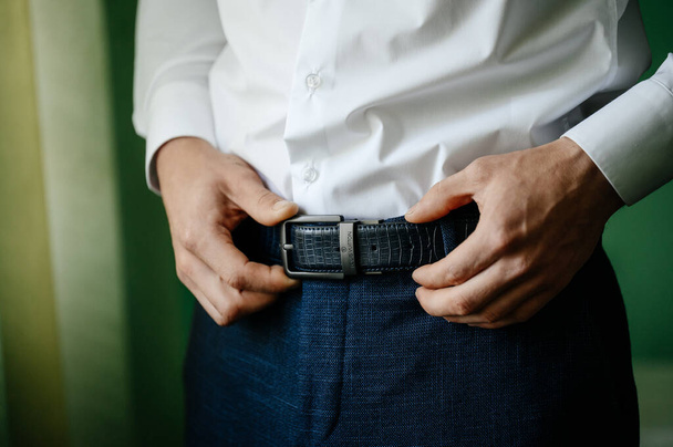 The morning of the groom, the groom is preparing for the wedding ceremony, dressing in a suit and adjusting the belt on his pants, place for inscription and copy space. - Фото, изображение