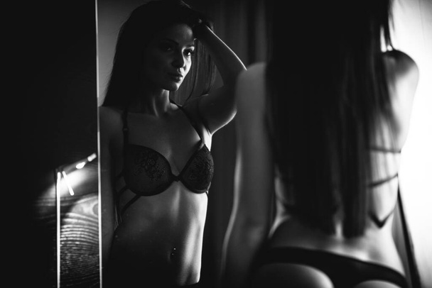Beauty of woman body and lingerie concept. Beautiful female model in sexy black underwear poses in luxury hotel room. Young girl stands by mirror wearing bra and panties. Black and white, monochrome. - Photo, Image