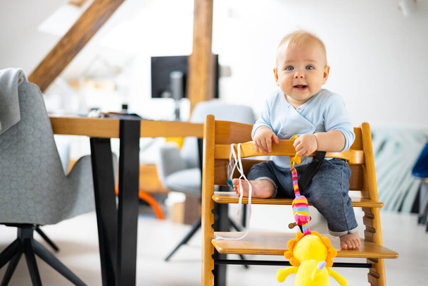Joyeux bébé assis et jouant avec son jouet dans une chaise haute en bois design scandinave traditionnelle dans une maison moderne et lumineuse. Bébé mignon - Photo, image