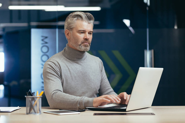 Serious and pensive mature investor working inside modern office, senior gray-haired businessman using laptop at work, male boss at work sitting at desk. - Foto, afbeelding