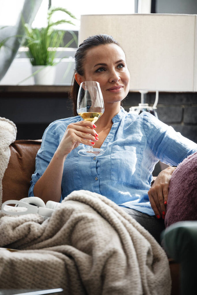 Leisure time concept. Happy beautiful woman drinks white wine from glass sitting on a couch indoors. Female spending her free day and relaxing at home alone - Photo, image