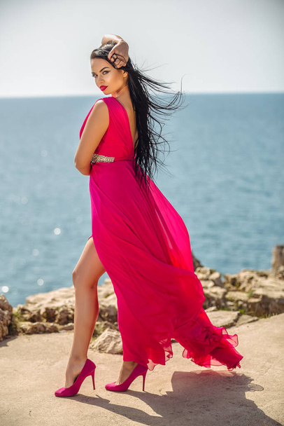 Beautiful black-haired girl in a red long dress on the seashore enjoys a summer day - Fotografie, Obrázek