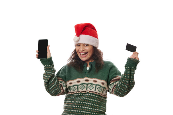 Uśmiechnięta radosna kobieta w ciepłym zielonym swetrze i kapeluszu Mikołaja, trzymająca w rękach czarną kartę kredytową i smartfona, odizolowana na białym tle. Bankowość internetowa, zakupy online, koncepcja aplikacji mobilnej - Zdjęcie, obraz