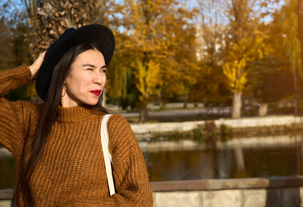 El retrato de cerca de una chica con un sombrero negro y un ojo de abrigo marrón guiña a la cámara y sonríe mientras sostiene la bolsa blanca. Enfoque selectivo y espacio de copia. Foto de alta calidad - Foto, imagen