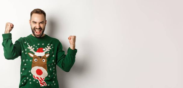 New Year, holidays and celebration. Excited bearded man in Christmas sweater, making fist pumps and shouting for joy, rejoicing and triumphing, white background. - Photo, Image