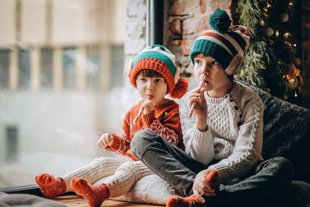 Δύο αδερφάκια κάθονται κοντά στο παράθυρο και περιμένουν τα Χριστούγεννα και τον Άγιο Βασίλη. Ευτυχισμένα παιδικά χρόνια και Χριστούγεννα. - Φωτογραφία, εικόνα