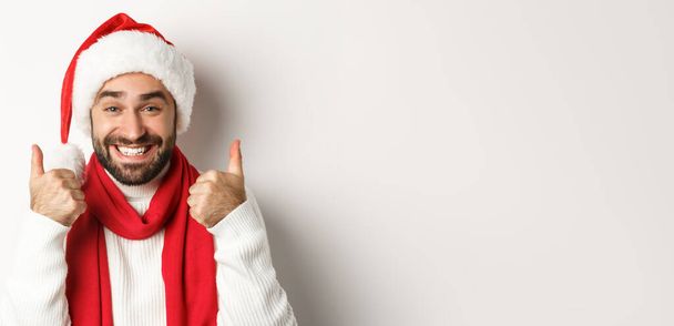 Novoroční párty a zimní prázdniny koncept. Detailní záběr šťastného muže v klobouku Santa ukazuje palce nahoru v souhlasu, jako a souhlasit, stojící nad bílým pozadím. - Fotografie, Obrázek