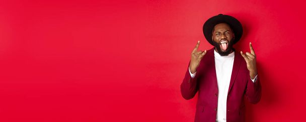 Szczęśliwy, afrykański mężczyzna bawiący się na imprezie, pokazujący język i rock-n-rollowy gest, świętujący Nowy Rok, stojący na czerwonym tle. - Zdjęcie, obraz