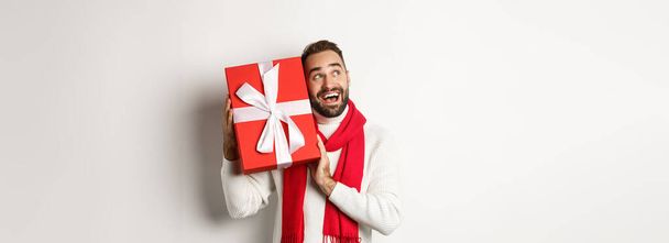 Концепция зимних каникул. Счастливый человек трясет подарочную коробку, чтобы угадать, что внутри, глядя любопытно, получать рождественские подарки, стоя на белом фоне. - Фото, изображение