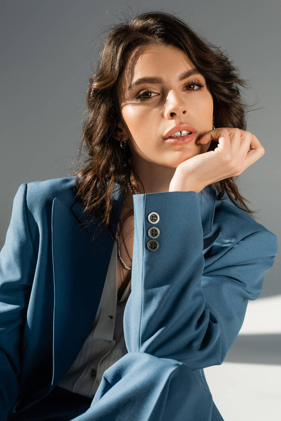 portrait of pretty brunette model in blue and stylish jacket looking at camera on grey background - Foto, Imagem