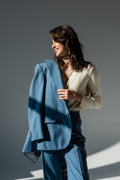 mujer feliz y elegante sosteniendo blazer azul mientras mira hacia otro lado sobre fondo gris - Foto, Imagen
