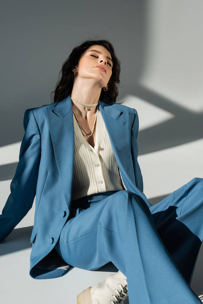trendy woman in blue suit sitting with closed eyes on grey background with lighting and shadows - Photo, Image