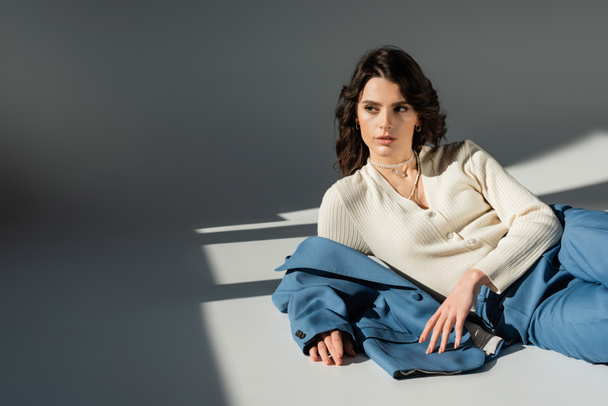 stylish brunette woman looking away while lying near blue jacket on grey background with lighting - Fotografie, Obrázek