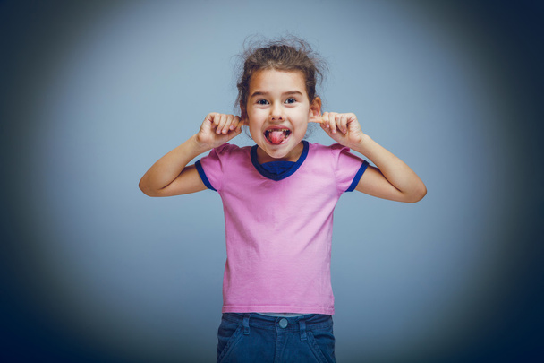 kind meisje weergegeven: tong trekt oren plaagt op een grijze achtergrond - Foto, afbeelding
