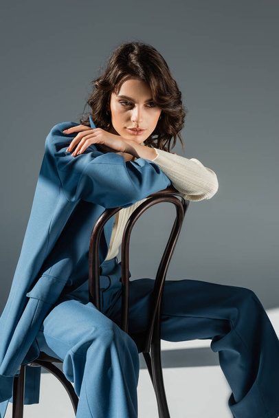 trendige brünette Frau sitzt auf Stuhl mit blauem Blazer auf grauem Hintergrund - Foto, Bild
