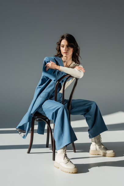full length of fashionable woman in pants and laced boots posing on chair near blue jacket on grey  - Foto, imagen