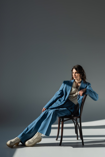 full length of stylish woman in blue suit and boots looking away on chair on grey background with lighting and shadows - Foto, imagen