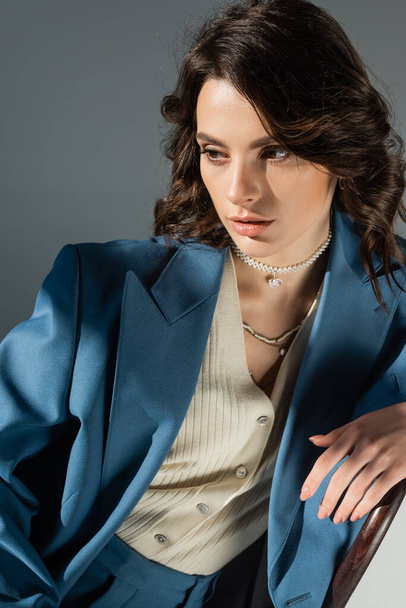 brunette woman in blue blazer and necklaces sitting and looking away on grey background - Fotoğraf, Görsel