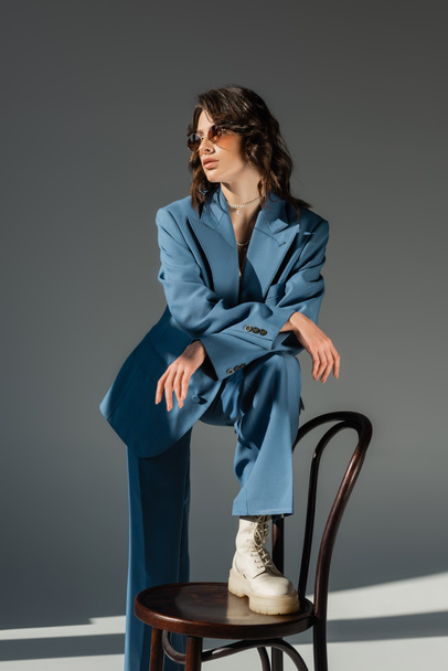 brunette woman in blue fashionable suit stepping on chair and looking away on grey background - Photo, Image