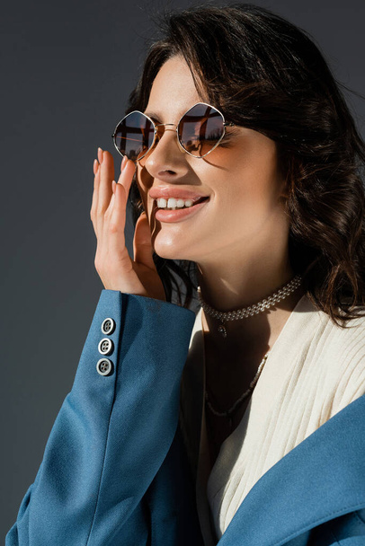 portrait of smiling fashionable woman in sunglasses holding hand near face isolated on grey - Photo, Image
