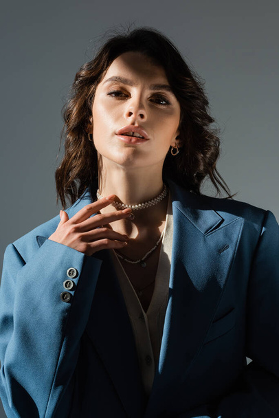чувственная брюнетка женщина в синей куртке трогает жемчужное ожерелье и смотрит на камеру, изолированную на сером - Фото, изображение