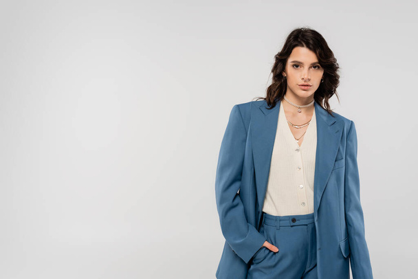 brunette woman in trendy blazer standing with hand in pocket of blue trousers isolated on grey - Foto, Imagem
