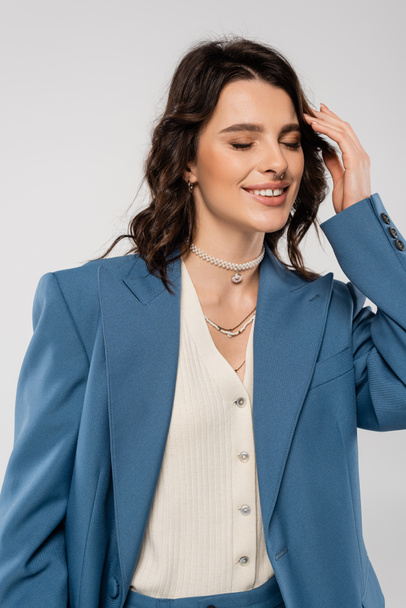 pleased woman in blue blazer and necklaces posing with closed eyes and touching hair isolated on grey - Foto, Imagen