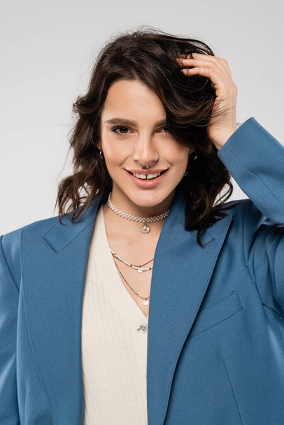 pretty woman with necklaces and piercing posing in blue blazer and smiling at camera isolated on grey - Photo, Image