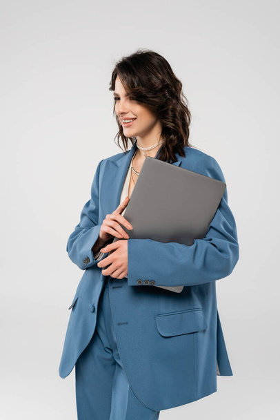 felice donna in giacca blu in piedi con computer portatile e guardando lontano isolato sul grigio - Foto, immagini
