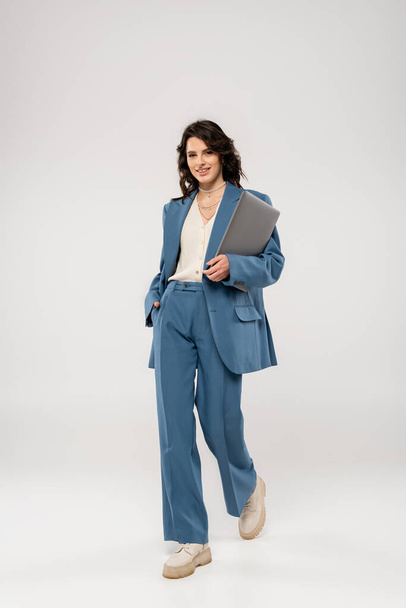 pełna długość radosnej kobiety w modnym garniturze trzymającej rękę w kieszeni podczas spaceru z laptopem na szarym tle - Zdjęcie, obraz