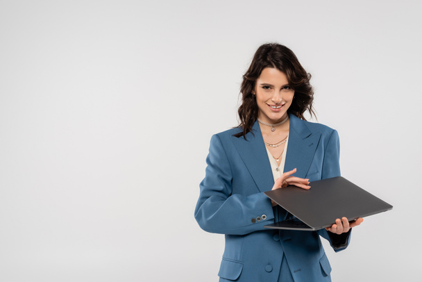 happy brunette woman in blue blazer holding laptop and looking at camera isolated on grey - Фото, изображение