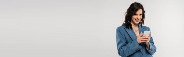 счастливая брюнетка женщина в синей куртке смотрит в камеру и сообщения на мобильный телефон изолированы на сером, баннер - Фото, изображение