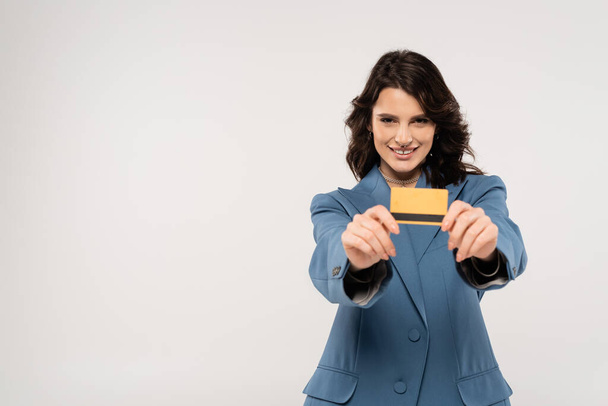 joyful brunette woman in blue jacket showing credit card at camera isolated on grey - Foto, Imagen