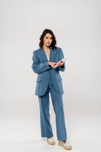 full length of trendy woman in blue suit using digital tablet and looking at camera on grey background - 写真・画像