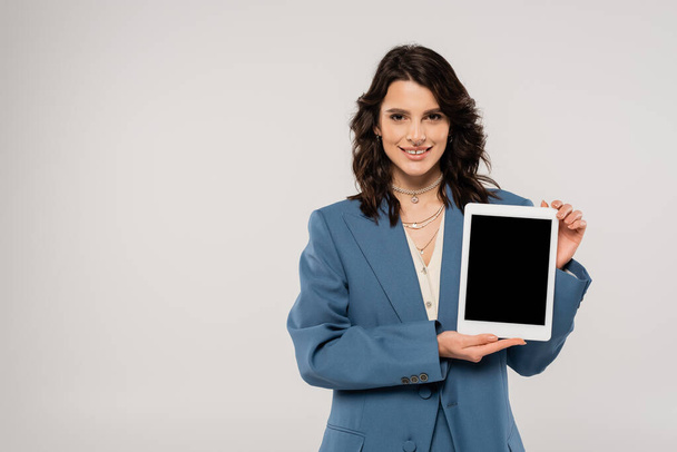 onnellinen brunette nainen sininen takki hymyilee kameralle ja näyttää digitaalinen tabletti tyhjä näyttö eristetty harmaa - Valokuva, kuva