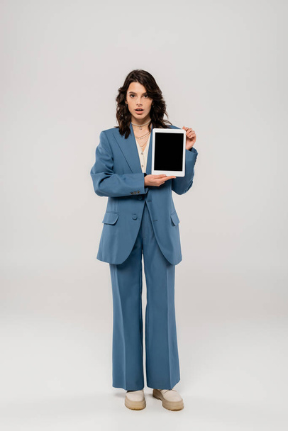plná délka překvapené ženy v modrém obleku zobrazující digitální tablet s prázdnou obrazovkou na šedé  - Fotografie, Obrázek