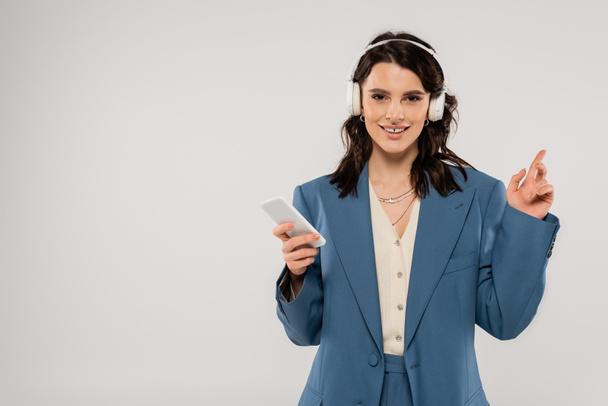 tyytyväinen brunette nainen langattomat kuulokkeet tilalla älypuhelin ja sormella eristetty harmaa - Valokuva, kuva